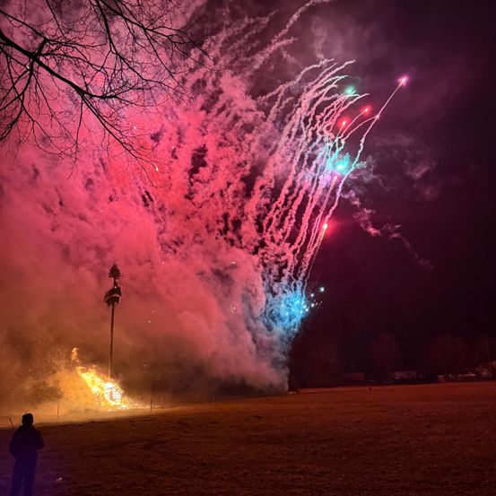 Ein farbenfroher Feuerwerksdisplay mit leuchtenden roten, blauen und grünen Farben, die einen dunklen Nachthimmel erhellen. Unten brennt ein Feuer mit einem hohen Pfahl in der Mitte.