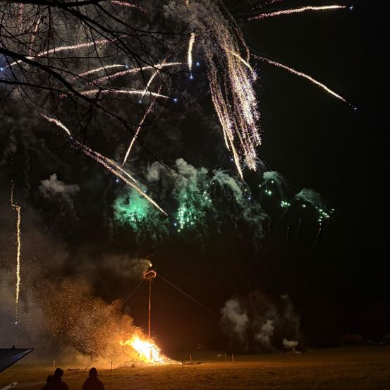 Ein nächtliches Feuerwerk mit leuchtenden Farben wie Grün und Weiß, das den dunklen Himmel erhellt. Ein Lagerfeuer brennt im Vordergrund, zwei Personen beobachten es.