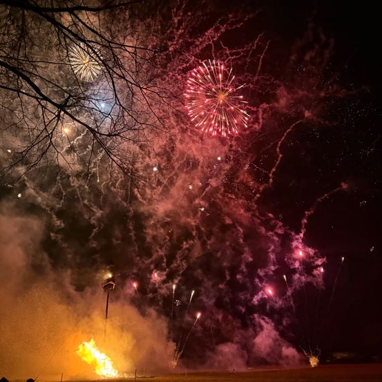 Ein Feuerwerk erleuchtet die Nacht mit roten und rosa Explosionen, ein zentrales Feuerwerk explodiert in der Mitte unten.