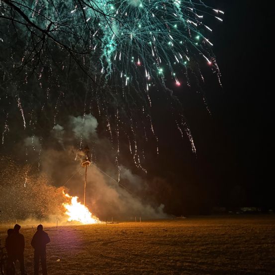 Ein Lagerfeuer mit Feuerwerken, die den Nachthimmel erhellen. Zwei Personen stehen in der Nähe des Feuers. Der Himmel ist dunkel, wobei das Feuer und die Feuerwerke die einzige Lichtquelle sind.