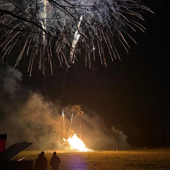 Ein nächtliches Feuerwerk mit Menschen, die auf einem Feld stehen, aufsteigendem Rauch und Bäumen im Vordergrund.