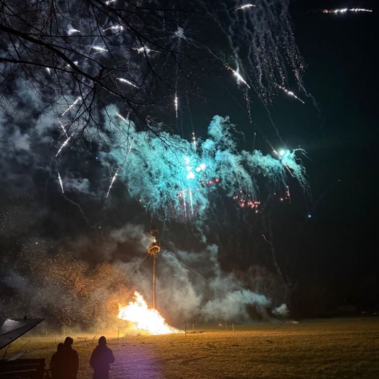 Ein Feuerwerk in einem Feld mit zwei Personen, die zuschauen. Die Show zeichnet sich durch leuchtende Farben und explodierende Feuerwerke am Nachthimmel aus.