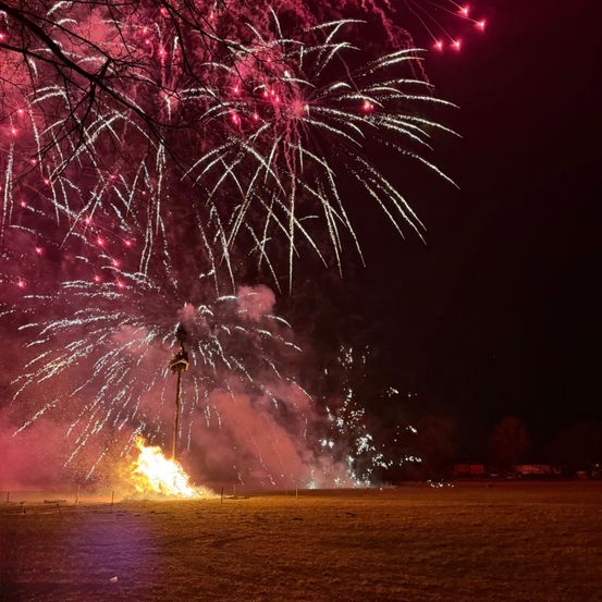 Ein Nachthimmel voller lebhafter Feuerwerke, die in verschiedenen Farben und Mustern gegen den dunklen Hintergrund explodieren. Ein Lagerfeuer brennt hell am Boden und sendet Funken nach oben.