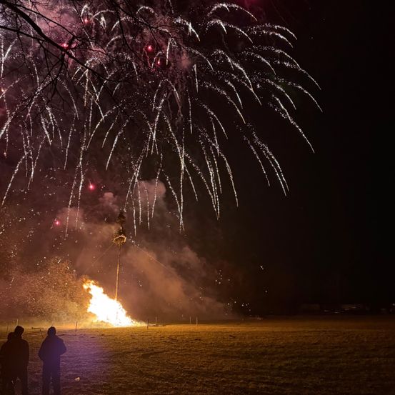 Eine nächtliche Szene mit einem Feuer, das unter einem Feuerwerksdisplay am Himmel brennt. Zwei Personen stehen im Vordergrund.