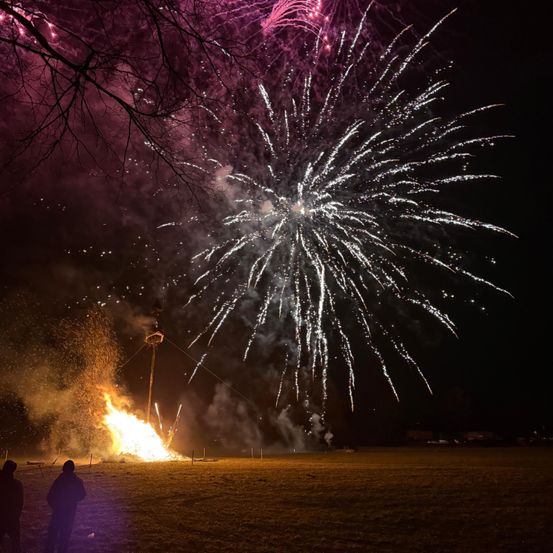 In der Nacht beleuchtet ein farbenfrohes Feuerwerk den Himmel, mit einem brennenden Lagerfeuer im Vordergrund. Bäume ohne Blätter umgeben das Gebiet.