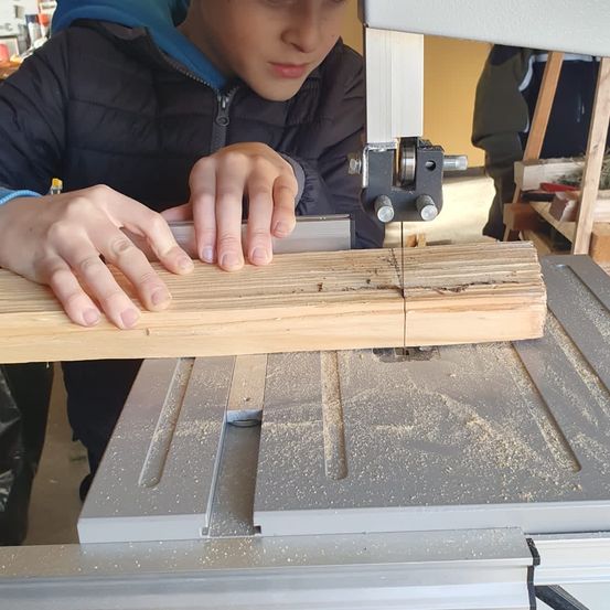 Ein junger Mensch schneidet mit einer Tischkreissäge ein Holzbrett, trägt eine blaue Jacke. Ein Holzregal ist im Hintergrund.