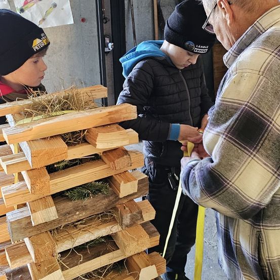 Drei Personen sind in einem Raum und arbeiten mit Holz. Einer beobachtet, während die anderen beiden an einem Stapel von Holzplanken arbeiten.