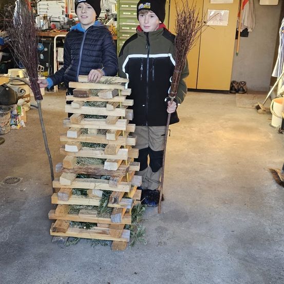 Zwei Jungen stehen in einer Garage, halten Besen in den Händen und posieren neben einem Stapel aus Holzpaletten, gefüllt mit Heu.
