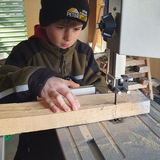 Ein junger Junge benutzt eine Tischkreissäge, um ein Stück Holz zu schneiden. Er trägt eine schwarze Mütze und eine Jacke mit reflektierenden Streifen.