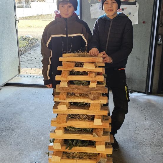 Zwei Jungen stehen neben einem hohen Stapel aus Holzpaletten, die mit Heu gefüllt sind. Einer trägt einen schwarzen und der andere einen braun-schwarzen Mantel. Sie lächeln und scheinen für ein Foto zu posieren.