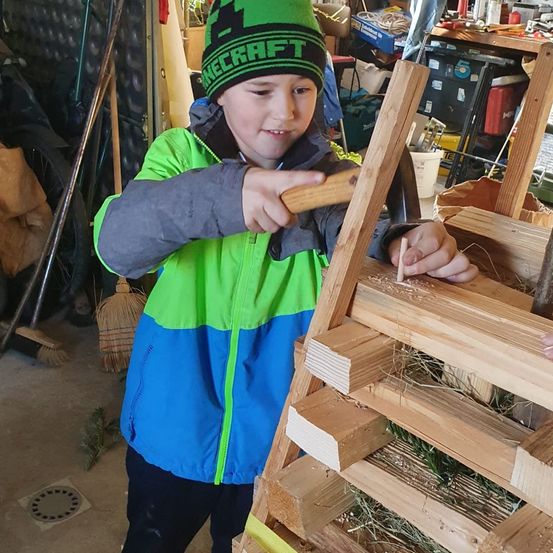 Ein junger Junge in einer grün-blauen Jacke und einer grünen Mütze baut mit einem Hammer an einer Holzstruktur. Der Schauplatz ist eine Garage mit verschiedenen Werkzeugen und Geräten.