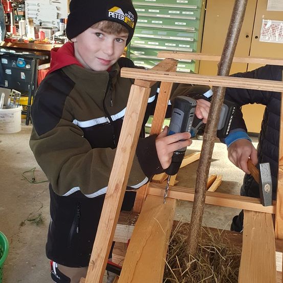 Ein junger Junge mit Mütze bohrt in eine Holzkonstruktion, während eine andere Person dahinter einen Hammer hält. Der Schauplatz scheint eine Werkstatt zu sein, mit verschiedenen Werkzeugen und Materialien im Hintergrund.
