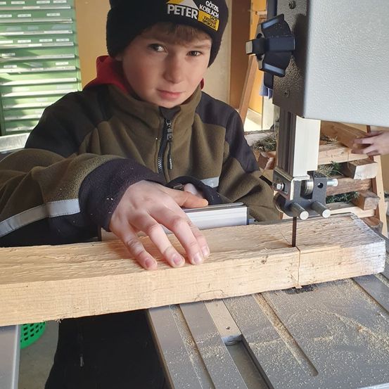 Ein junger Junge in einer Mütze und einer Jacke säg einen Holzbalken mit einer Bandsäge.