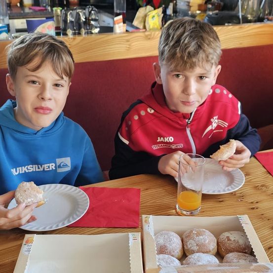 Zwei Jungen sitzen an einem Tisch, einer in einer blauen Kapuzenjacke und der andere in einer roten Jacke, beide essen Donuts. Ein Glas Orangensaft und eine Schachtel Donuts stehen auf dem Tisch.