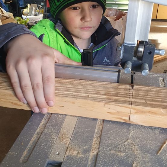 Ein junger Junge in einem grünen Weste und Mütze benutzt eine Tischkreissäge, um ein Stück Holz zu schneiden. Das Holz wird von der Säge gehalten.