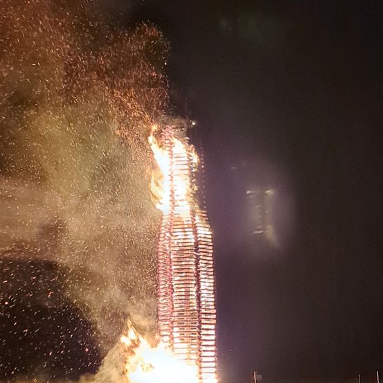 Ein hoher Holzbau brennt nachts hell, Flammen und Funken erhellen den dunklen Himmel.