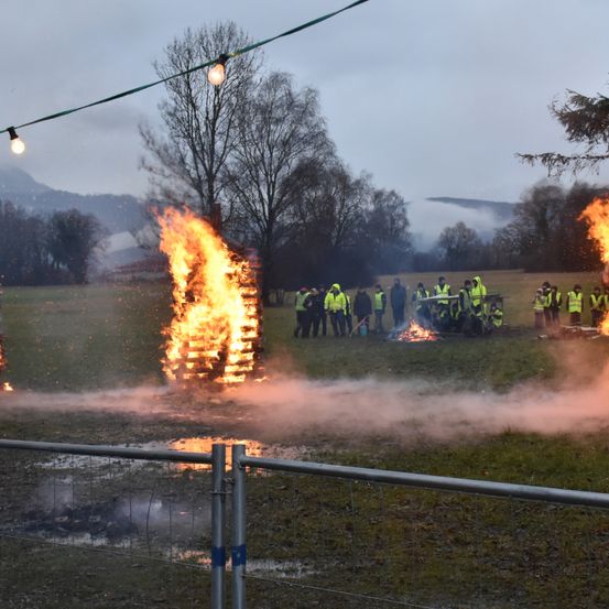 Eine Gruppe von Menschen in gelben Westen steht um zwei große Lagerfeuer in einem Feld, mit Lichterketten und Bäumen im Hintergrund.