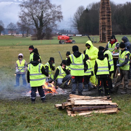 Eine Gruppe von Menschen in neongrünen Warnwesten steht um ein brennendes Feuer auf einem Feld, mit einem großen Holzstapel im Hintergrund.
