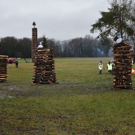 Eine Gruppe von Menschen bereitet sich darauf vor, drei große Holzstrukturen mit Bäumen auf der Oberseite auf einer Wiese zu verbrennen.