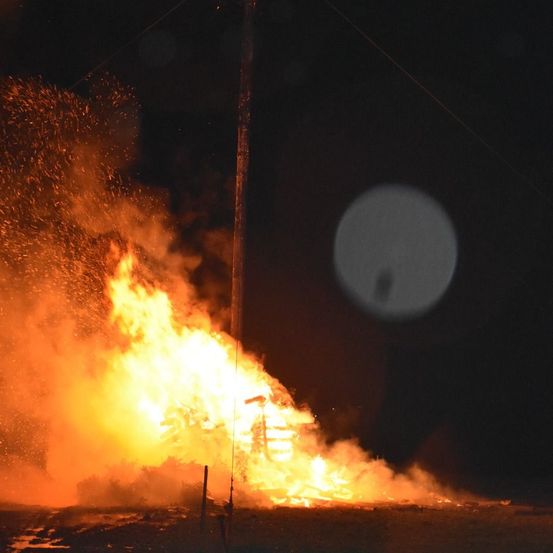 Ein großes Lagerfeuer brennt bei Nacht hell mit fliegenden Funken, nahe einem Pfosten und einem mysteriösen Licht.