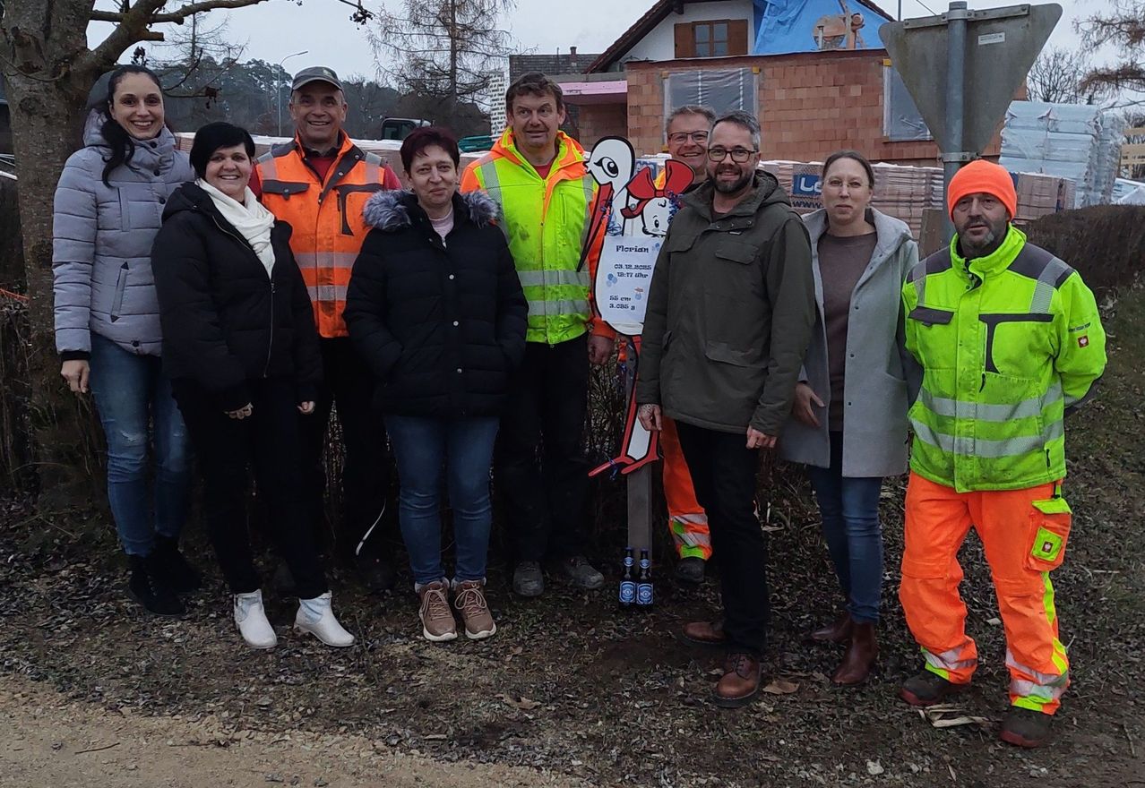 Eine Gruppe von Menschen in Winterkleidung, einschließlich Warnwesten, steht vor einem im Bau befindlichen Gebäude. Ein Schild mit einer Comic-Figur ist vorhanden.