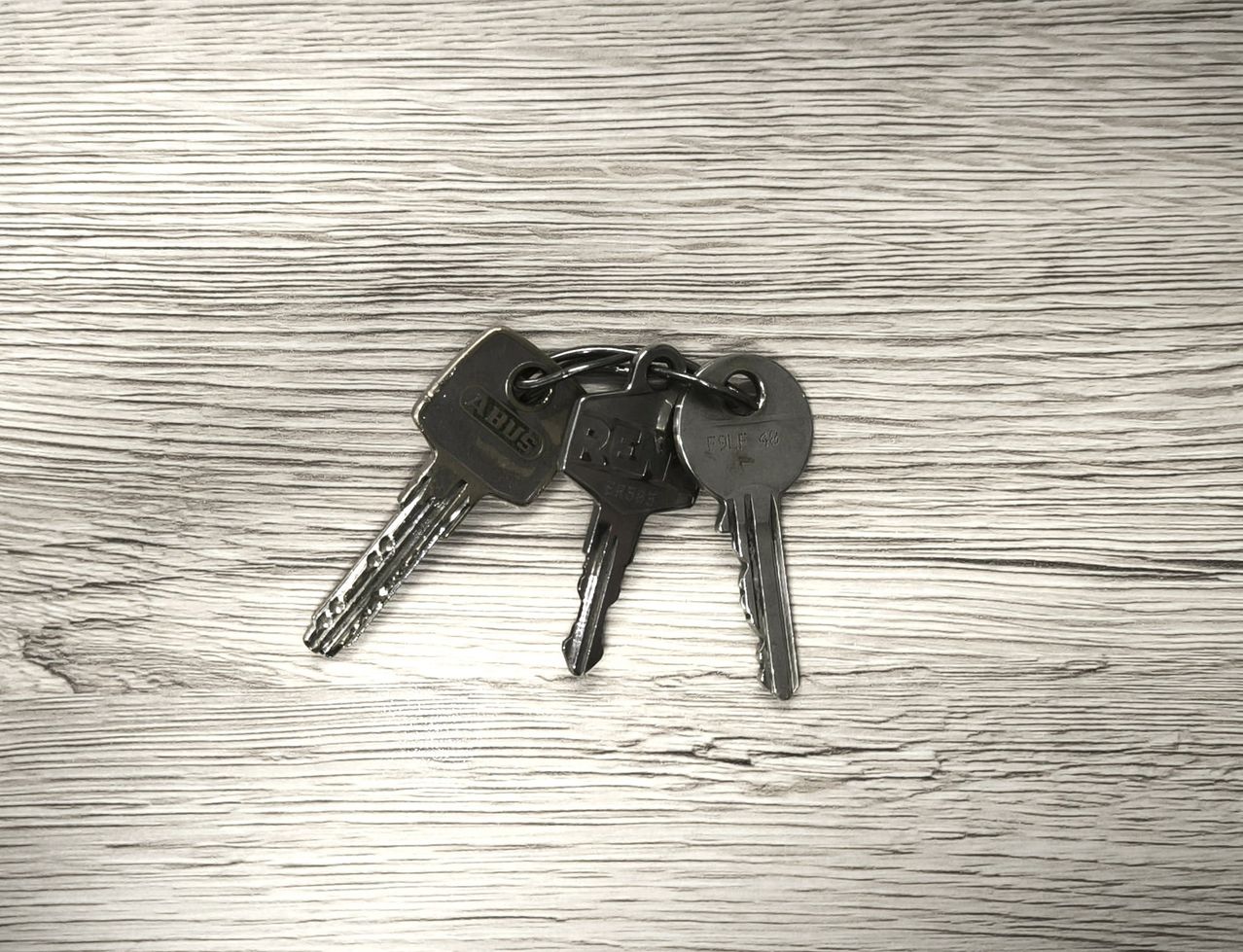 Drei schwarze Schlüssel mit den Aufschriften 'ABIS', 'RH' und 'FOLP 40' liegen auf einer Holzoberfläche. Die Schlüssel erscheinen metallisch und haben einen kleinen Schlüsselanhänger.