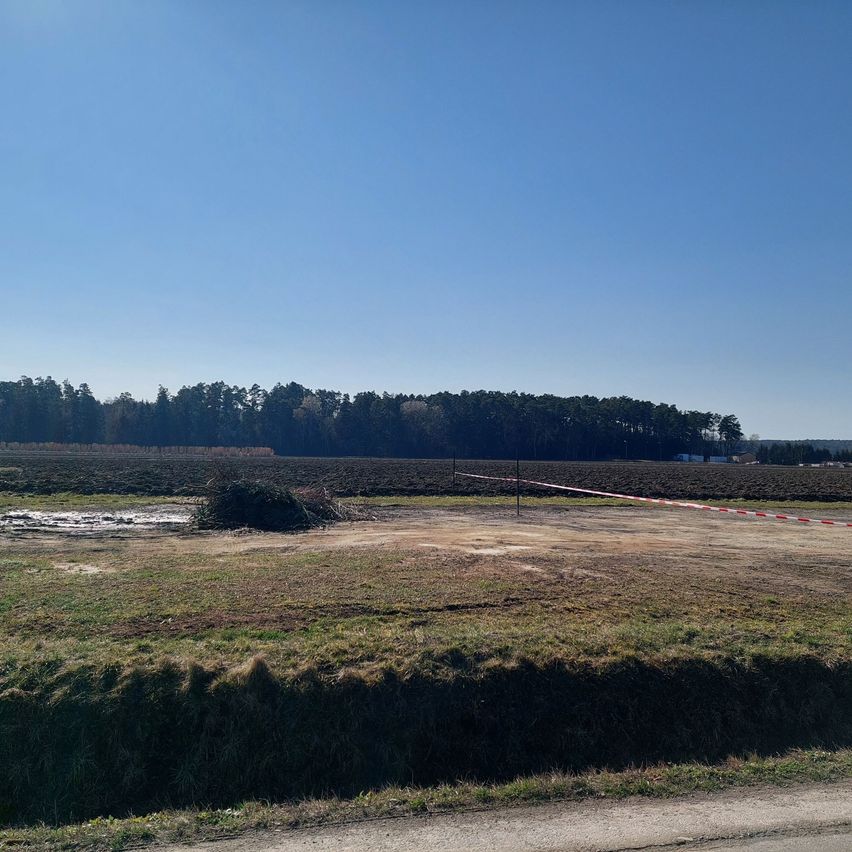 Ein offenes Feld mit Gras und einem kleinen Teich. Ein Zaun mit rot-weißen Streifen blockiert die Mitte. Bäume sind im Hintergrund unter einem klaren blauen Himmel sichtbar.