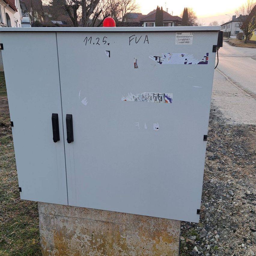 Ein grauer Versorgungskasten mit zwei Türen, Graffiti und Aufklebern, der auf einem Betonsockel im Freien steht.