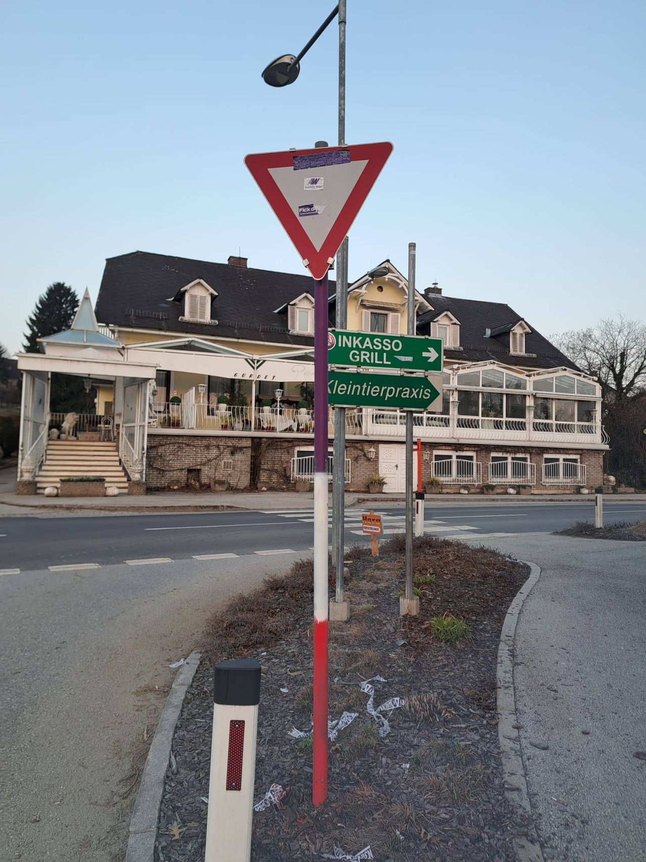 Eine Straßenecke mit einem dreieckigen Schild und zwei grünen Straßenschildern. Das Gebäude dahinter hat ein schwarzes Dach und Glasfenster. Es gibt eine Treppe, die zum Eingang führt.