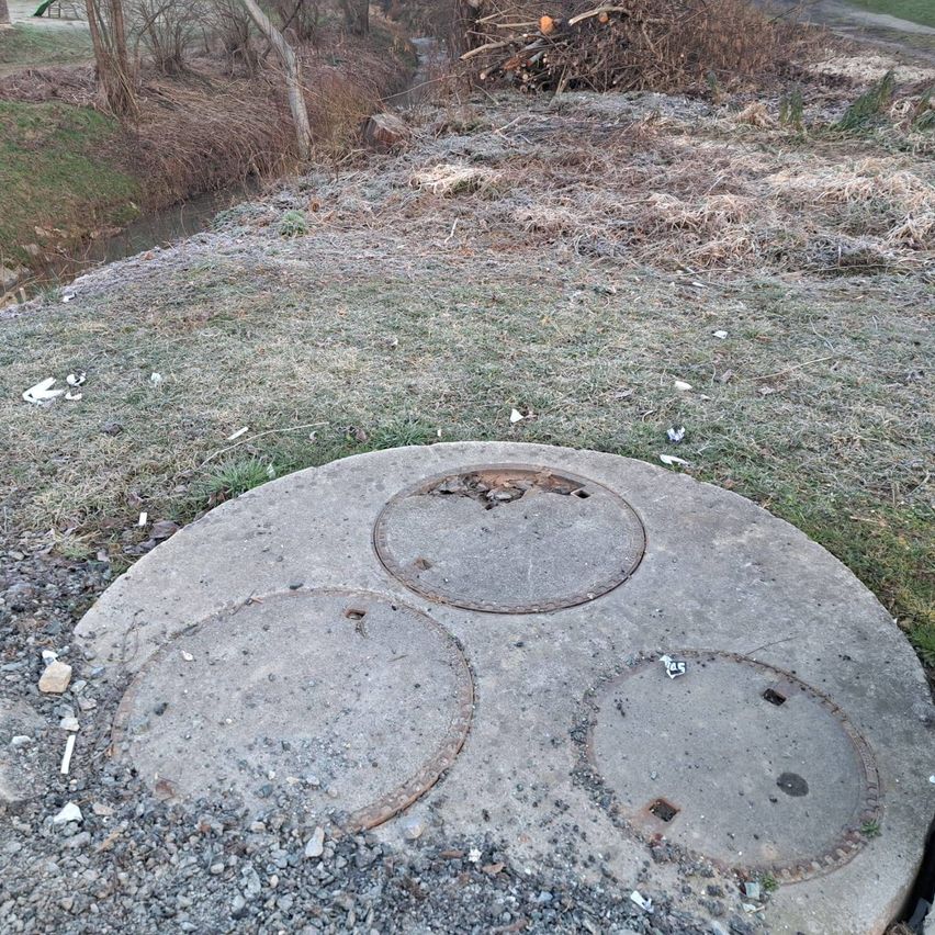 Drei runde Betonschachtdeckel liegen auf dem Boden mit Schutt. Hinter den Deckeln befindet sich ein Grasbereich mit einem kleinen Wasserlauf.