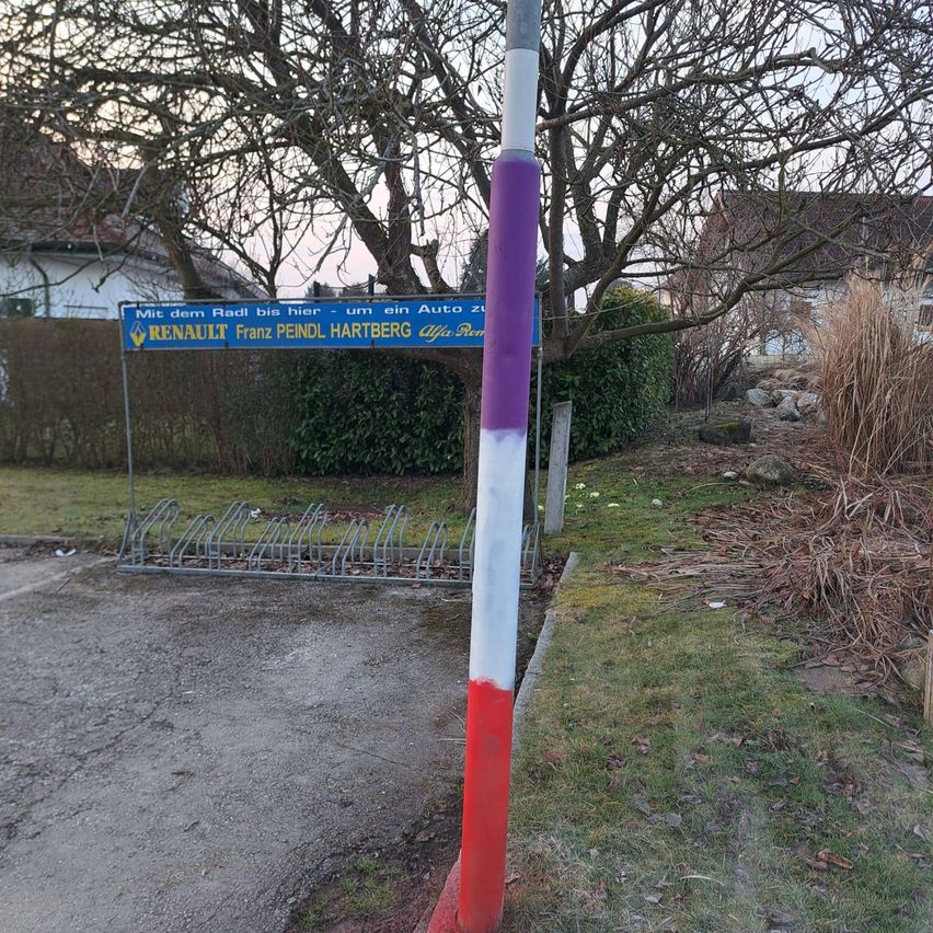 Ein rot, weiß und lila gestrichener Pfosten steht auf einem Parkplatz. Ein blaues Schild ist an einem Metallständer hinter dem Pfosten befestigt. Ein Haus mit braunem Dach befindet sich hinter einem Busch.