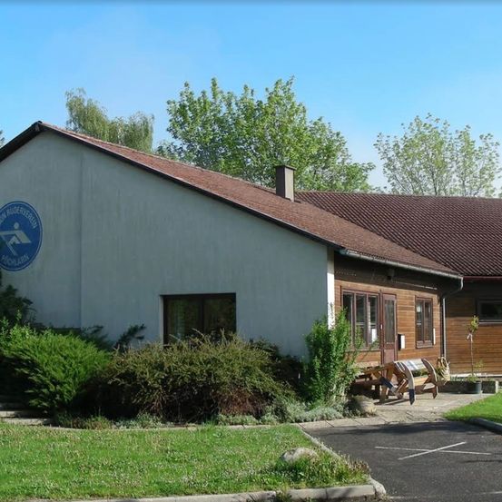 Ein Gebäude mit einem braunen Dach und einem Logo an der Wand. Es gibt Sträucher und Bäume um das Gebäude herum und eine Bank davor.