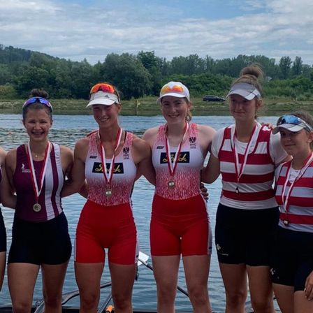 Fünf Frauen in Sportkleidung posieren für ein Foto am See. Sie tragen Medaillen und Mützen. Dahinter befindet sich ein bewaldetes Gebiet und ein klarer Himmel.