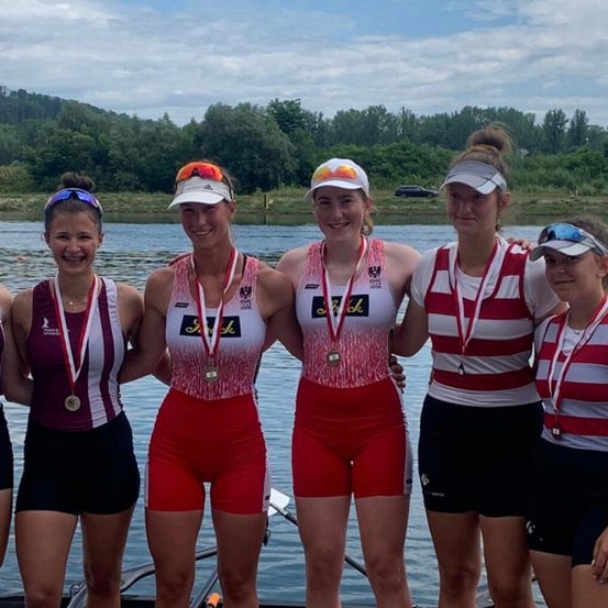 Fünf Frauen in Sportkleidung posieren für ein Foto am See. Sie tragen Medaillen und Mützen. Dahinter befindet sich ein bewaldetes Gebiet und ein klarer Himmel.