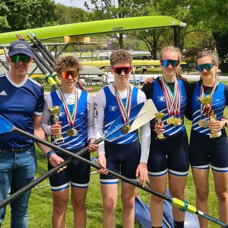 Eine Gruppe von fünf Personen, wahrscheinlich ein Ruderteam, steht auf einem Rasenfeld. Sie halten Pokale und Medaillen und tragen passende blaue Westen und Shorts. Ein Mann steht links und hält ein Paddel. Im Hintergrund sind Boote abgestellt.