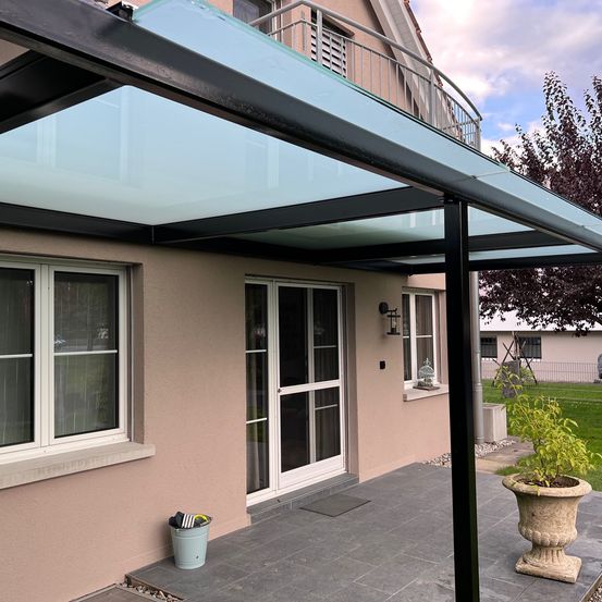 Ein modernes Haus mit einer Glasüberdachung über dem Eingang, einem Balkon mit Geländer und einer Terrasse mit einer Pflanze in einem Steintopf.