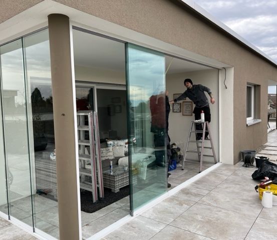Eine Person arbeitet an einer Schiebetür aus Glas. Eine Leiter und ein Eimer sind in der Nähe. Die Terrasse hat eine Fliesenboden.