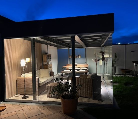 Ein modernes Glaszimmer mit einer Couch, einem Beistelltisch, einer Pflanze und einer Lampe am Abend.
