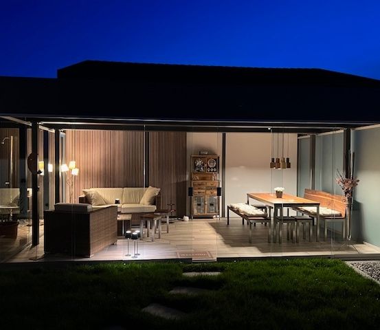 Ein moderner Wohnbereich im Freien mit einer Glaswand, einem Sofa, einem Tisch und einer Bank. Der Raum wird von Lampen beleuchtet, und der Boden ist gefliest.