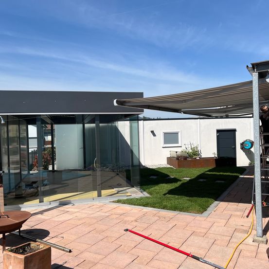 Eine moderne Terrassenüberdachung mit Glaswänden unter einem ausziehbaren Sonnensegel, mit einem Mann, der daran arbeitet.