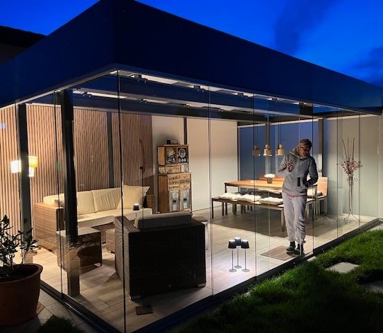 Ein modernes Glashaus mit Holzpaneelen und Glaswänden. Drinnen gibt es eine Couch, einen Tisch und eine Bank. Eine Person steht in der Ecke und ein Topf mit Pflanzen steht links.