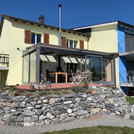 Ein modernes Haus mit einer Glaserweiterung, einer Terrasse und einer Steinmauer. Das Haus hat ein blau-gelbes Äußeres mit einer Terrasse, die mit einem Tisch und Stühlen ausgestattet ist.
