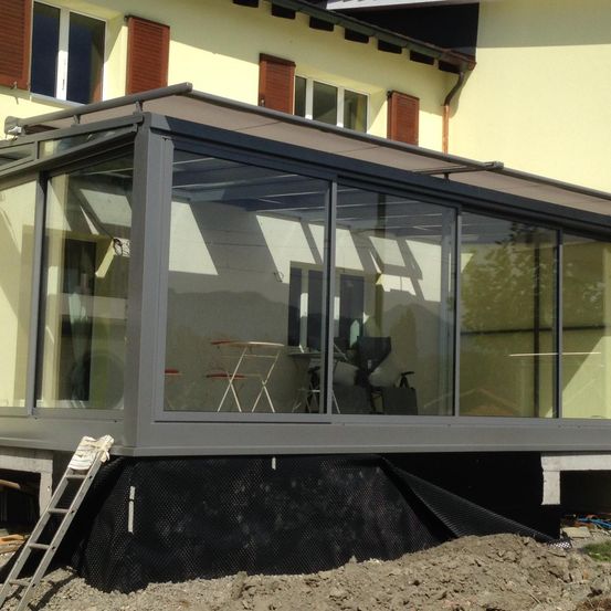 Eine Glasveranda mit Metallrahmen und Vordach, angeschlossen an ein gelbes Haus. Im Inneren befinden sich ein Tisch, Stühle und ein Grill. Der Außenbereich befindet sich im Bau.