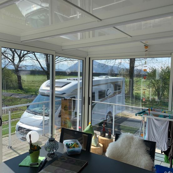 Eine Innenansicht eines Wohnmobils, das draußen geparkt ist. Es gibt Glastüren und -fenster, einen Balkon mit Geländern und einen Tisch mit verschiedenen Gegenständen. Der Blick nach draußen zeigt ein Grasfeld und Berge.