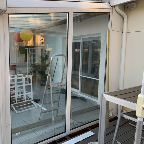 Eine Glasschiebetür ist geöffnet und reflektiert einen Innenraum mit einer Leiter, einer Pflanze in einem Topf und zwei hängenden Laternen. Ein Tisch und ein Stuhl sind rechts sichtbar.