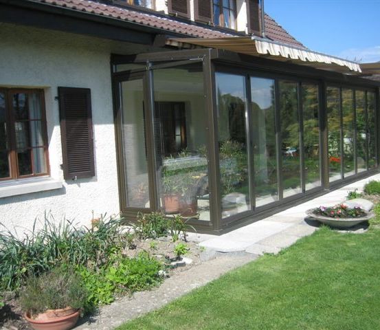 Ein weißes Haus mit einem braunen Dach hat einen Glasanbau mit Pflanzen und Blumen. Der Anbau hat ein weißes Vordach und ist von einem Garten mit verschiedenen Pflanzen umgeben.