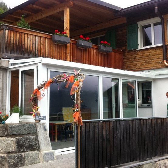 Ein Haus mit einem hölzernen Balkon, der mit roten Blumen geschmückt ist, ein Mann in Grün steht vor einer Glastür, und ein Kranz mit orangefarbenen Bändern hängt in der Nähe.