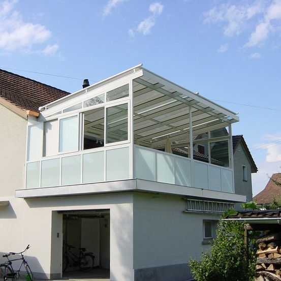 Ein weißes Haus mit einem glasüberdachten Balkon und einer Garage, Fahrräder vor dem Haus, unter einem teilweise bewölkten Himmel.