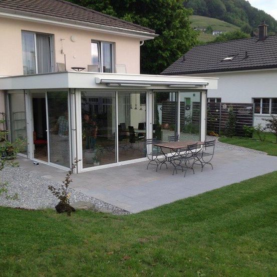Ein modernes Haus mit Terrasse und Glaswänden, neben einem kleineren Haus mit schwarzem Dach und Garten.