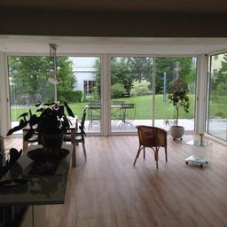 Ein offenes modernes Wohnzimmer mit Glaswänden und Türen, die zu einer Terrasse führen. Topfpflanzen, ein Esstisch und Stühle sind im Inneren, mit einem Mann, der sich neben den Glastüren bewegt.
